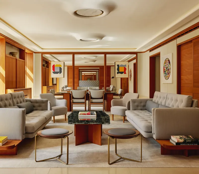 Contemporary living room in The Apartment with pale grey tufted sofas, armchairs, and a marble-topped coffee table, decorated with abstract art and wood paneling.