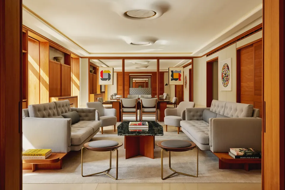 Contemporary living room in The Apartment with pale grey tufted sofas, armchairs, and a marble-topped coffee table, decorated with abstract art and wood paneling.