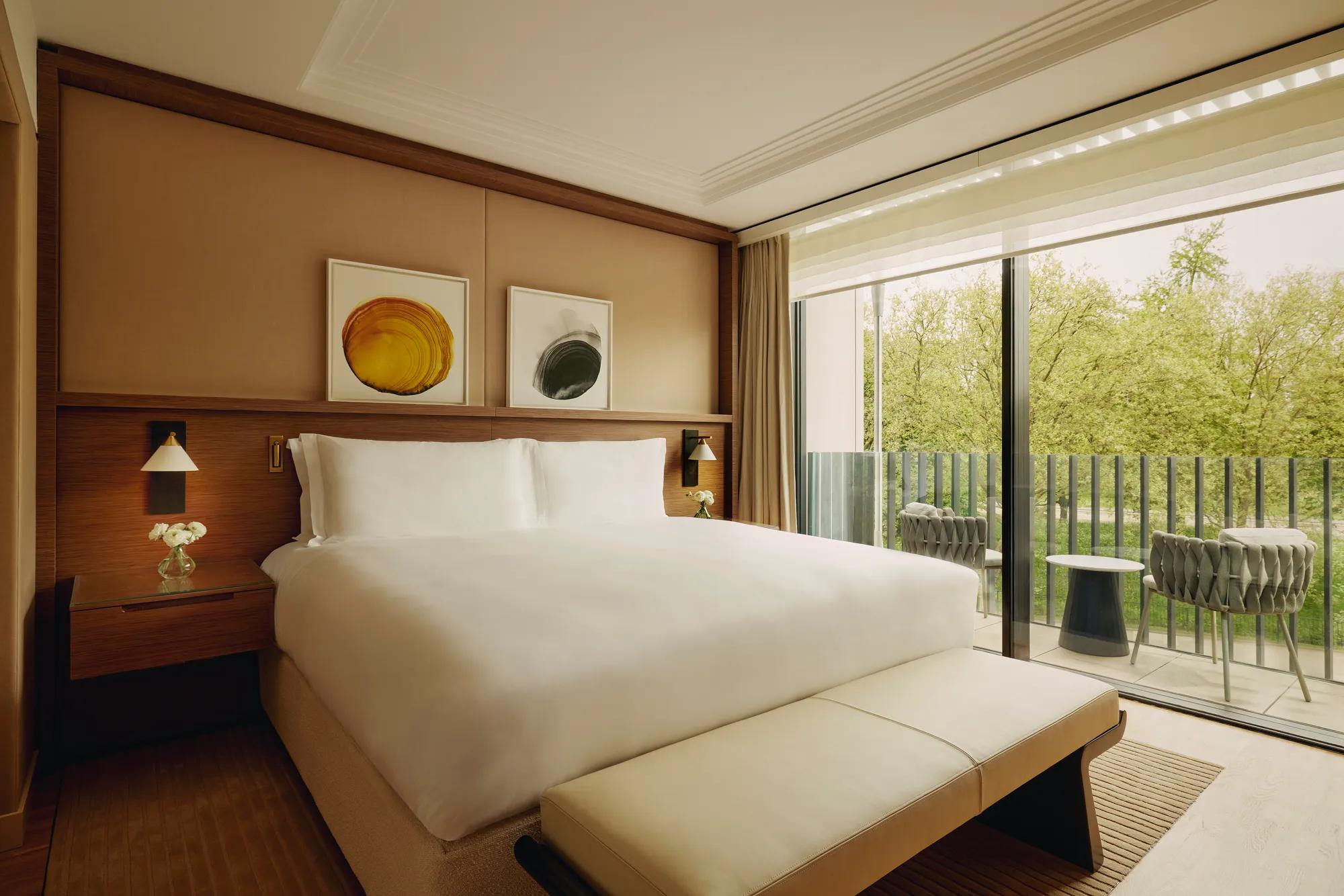 Elegant hotel bedroom with a large bed, modern artwork above the headboard, and floor-to-ceiling windows opening to a balcony with chairs and views of green trees.