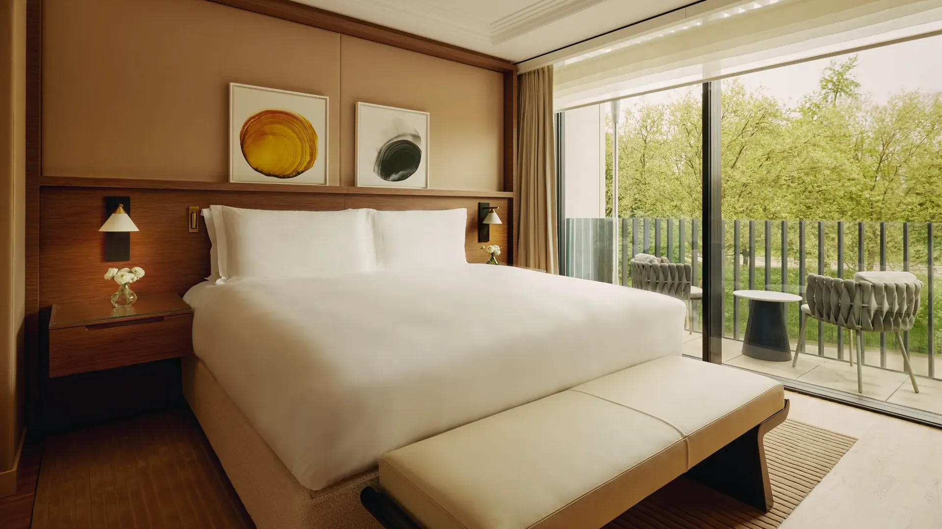 Elegant hotel bedroom with a large bed, modern artwork above the headboard, and floor-to-ceiling windows opening to a balcony with chairs and views of green trees.