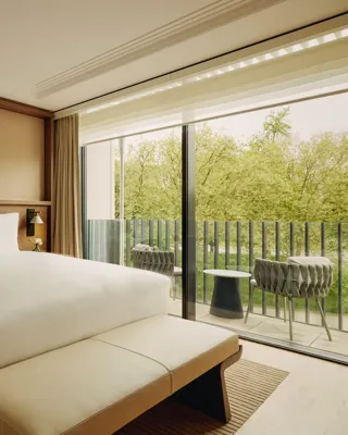 Elegant hotel bedroom with a large bed, modern artwork above the headboard, and floor-to-ceiling windows opening to a balcony with chairs and views of green trees.