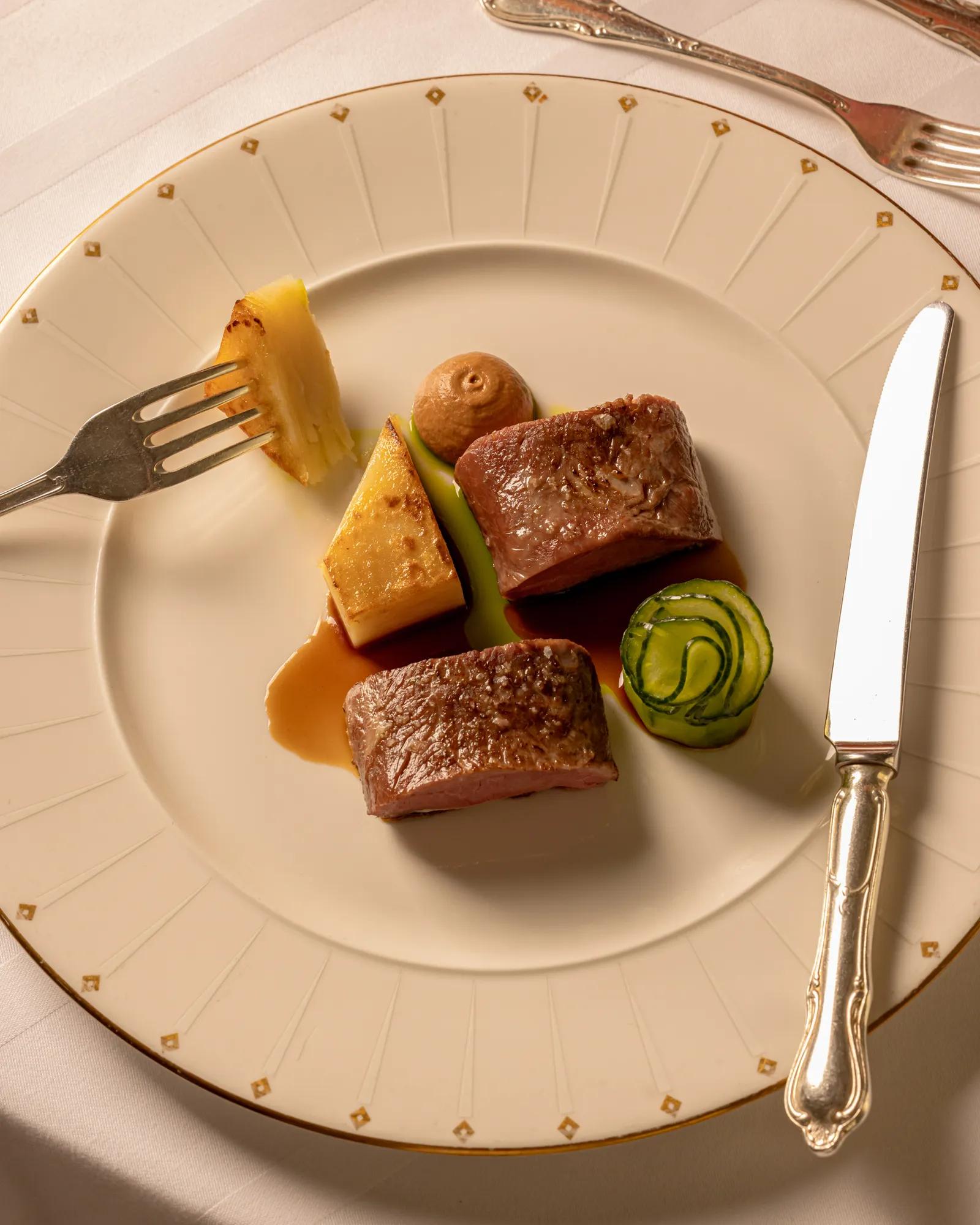 Fine dining dish presented on an elegant cream and gold-rimmed plate, featuring two pieces of seared meat with artfully arranged vegetables, sauce, and a small purée swirl, set with silver cutlery on a white tablecloth.