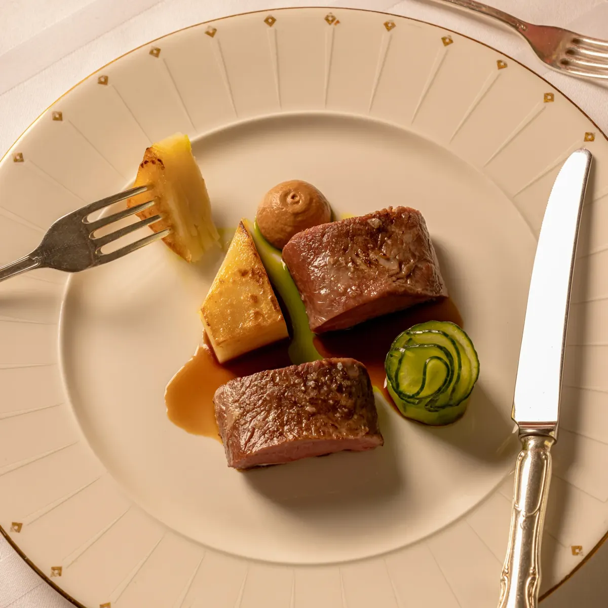 Fine dining dish presented on an elegant cream and gold-rimmed plate, featuring two pieces of seared meat with artfully arranged vegetables, sauce, and a small purée swirl, set with silver cutlery on a white tablecloth.