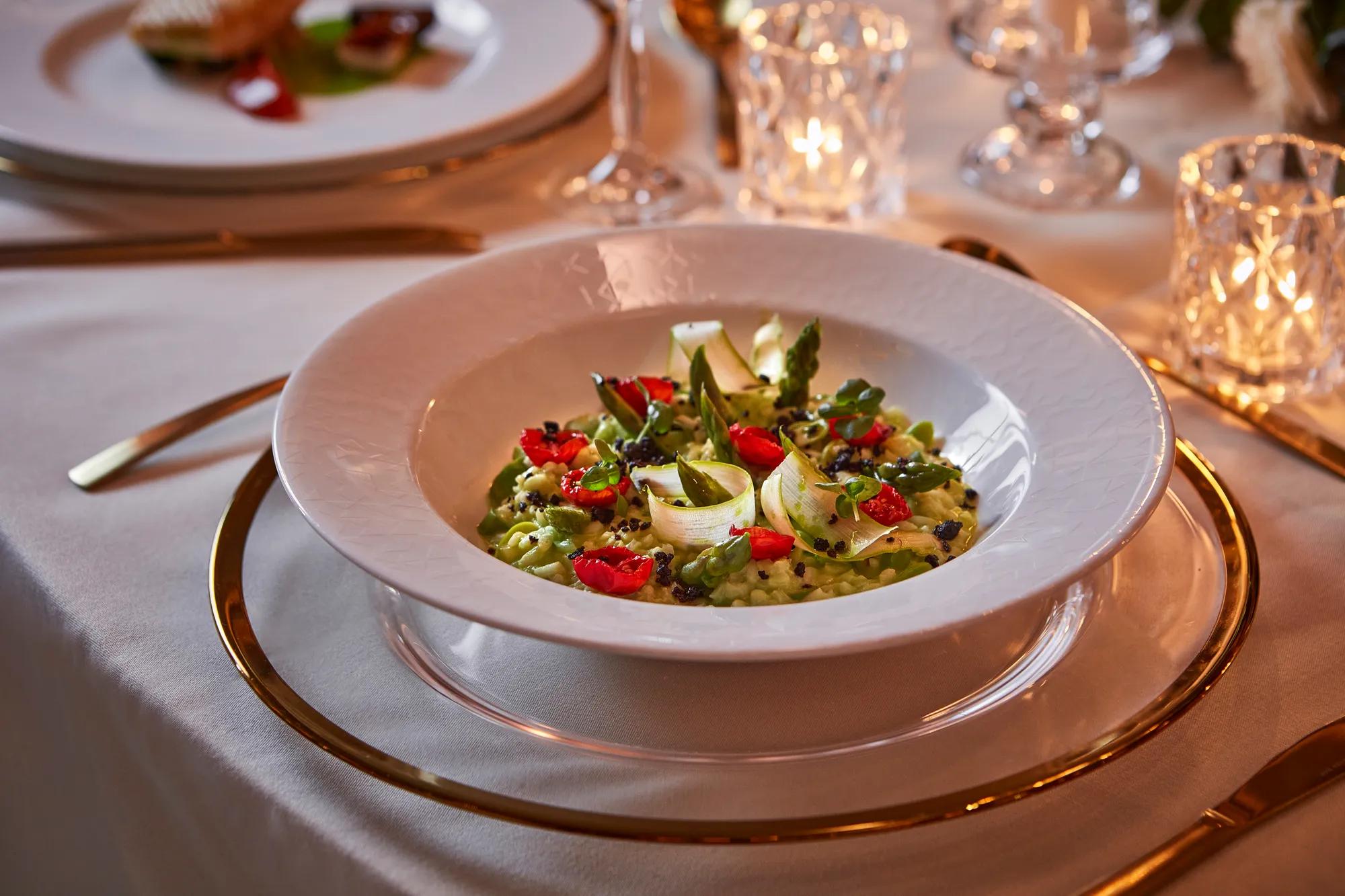 White bowl of green risotto with asparagus, tomato, and microgreens, served on a gold-rimmed plate with candlelit table setting.