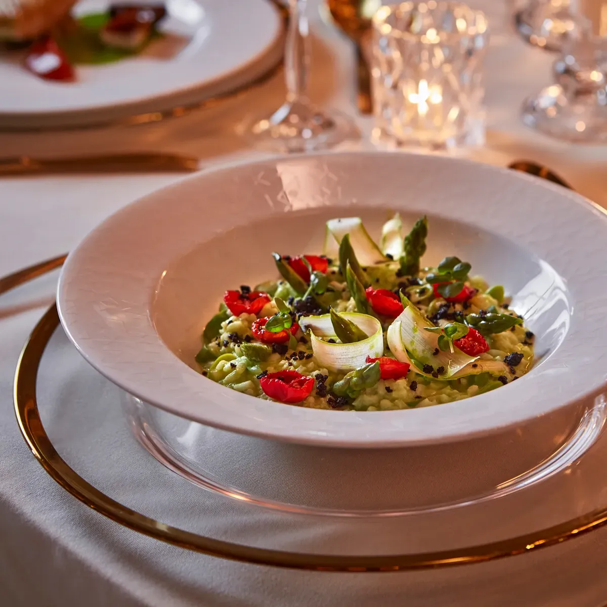 White bowl of green risotto with asparagus, tomato, and microgreens, served on a gold-rimmed plate with candlelit table setting.