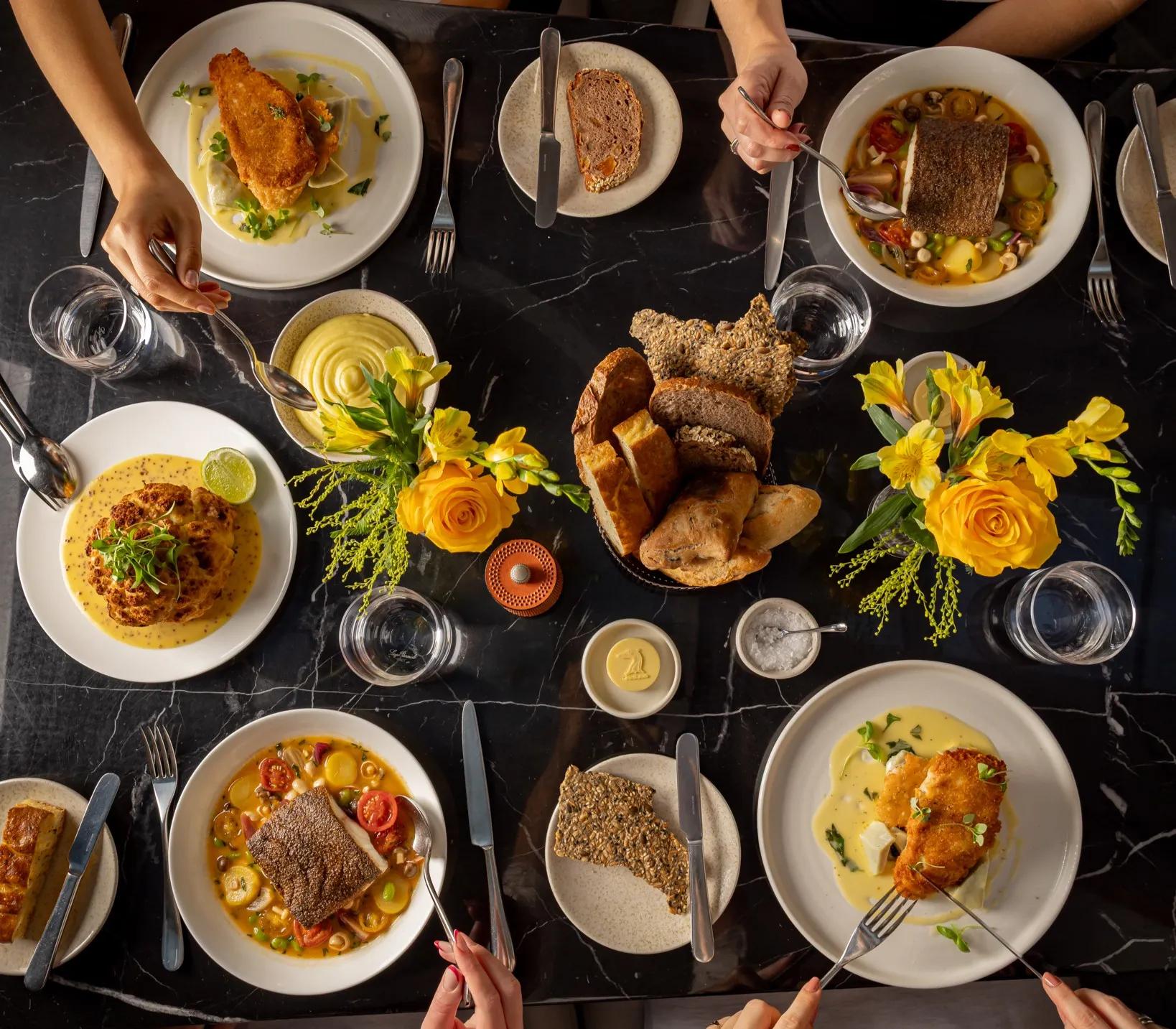 Overhead view of a shared dining table with plated mains, bread, yellow floral arrangements, and guests’ hands serving food.