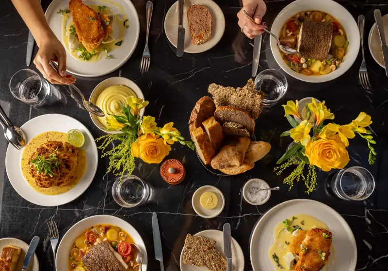 Overhead view of a shared dining table with plated mains, bread, yellow floral arrangements, and guests’ hands serving food.