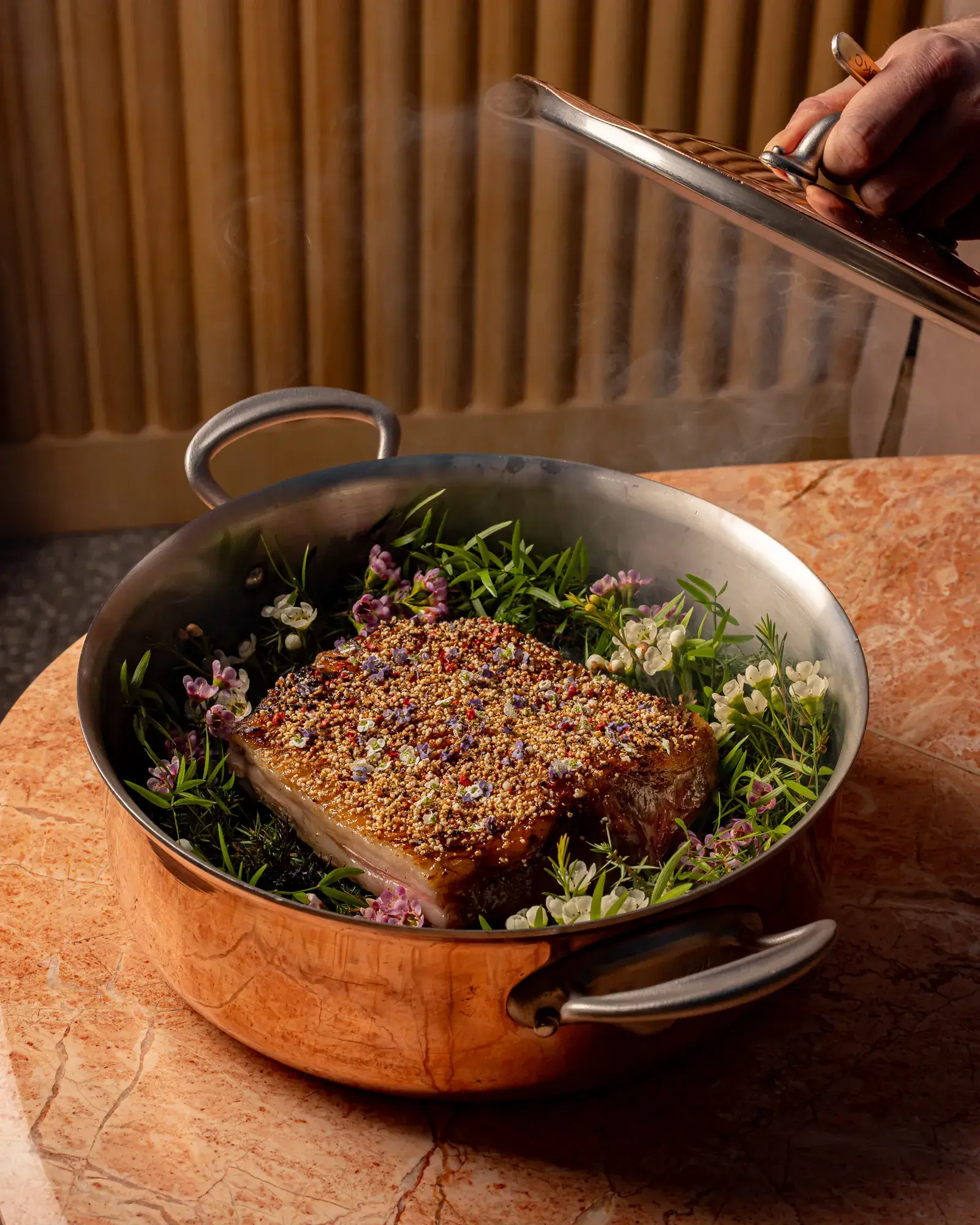 Chef lifting lid from copper pan revealing herb-crusted meat resting on fresh herbs and flowers, with steam rising.