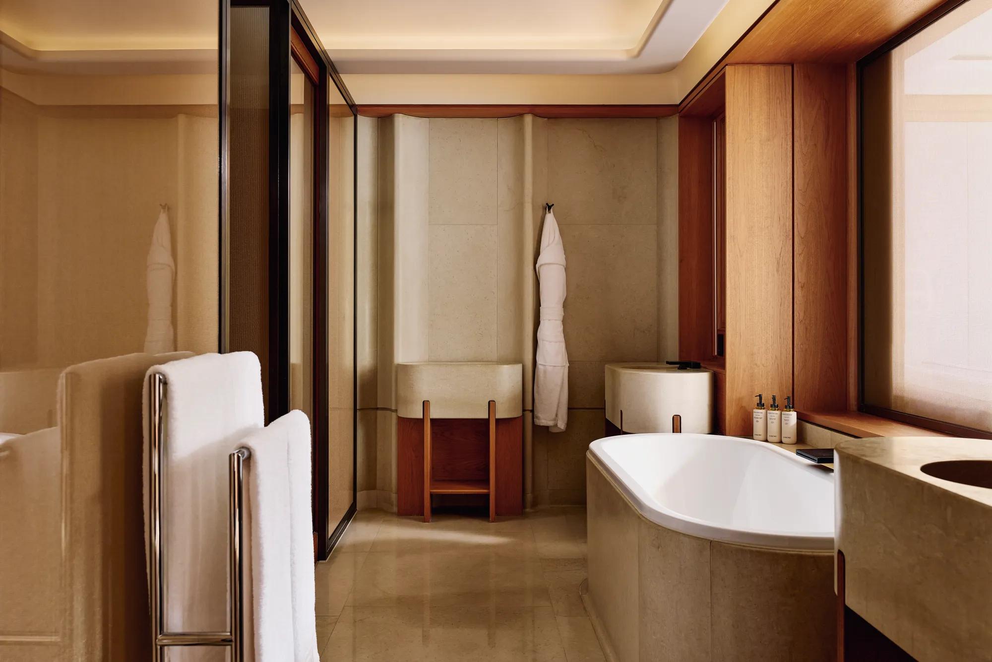 A modern bathroom with beige stone walls and flooring, featuring a deep bathtub, double sinks, and neatly arranged toiletries on the windowsill.