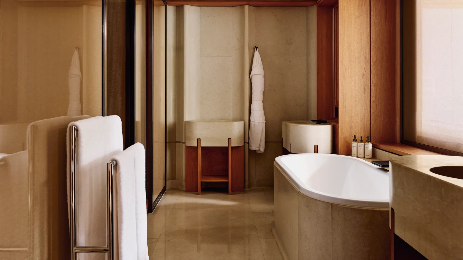 A modern bathroom with beige stone walls and flooring, featuring a deep bathtub, double sinks, and neatly arranged toiletries on the windowsill.