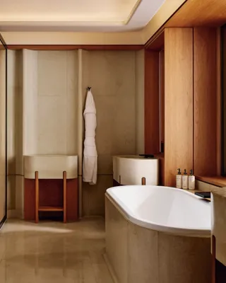A modern bathroom with beige stone walls and flooring, featuring a deep bathtub, double sinks, and neatly arranged toiletries on the windowsill.