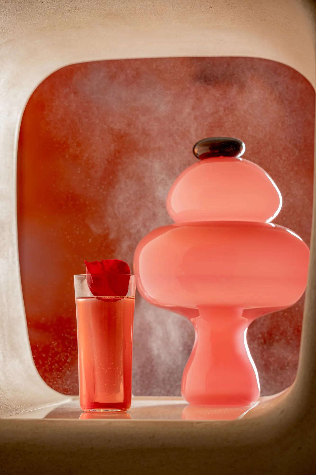 Pink cocktail garnished with a rose petal beside a sculptural pink lamp, displayed in an illuminated niche with a soft red backdrop.