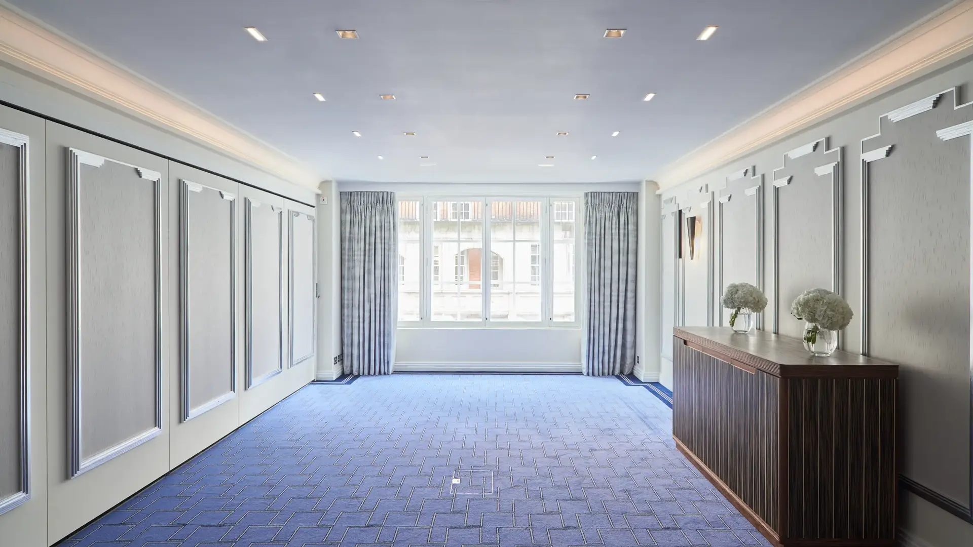 Bright, modern event room at Claridge’s featuring blue patterned carpet, soft grey panelled walls, large curtained windows, and a wooden sideboard decorated with white floral arrangements.