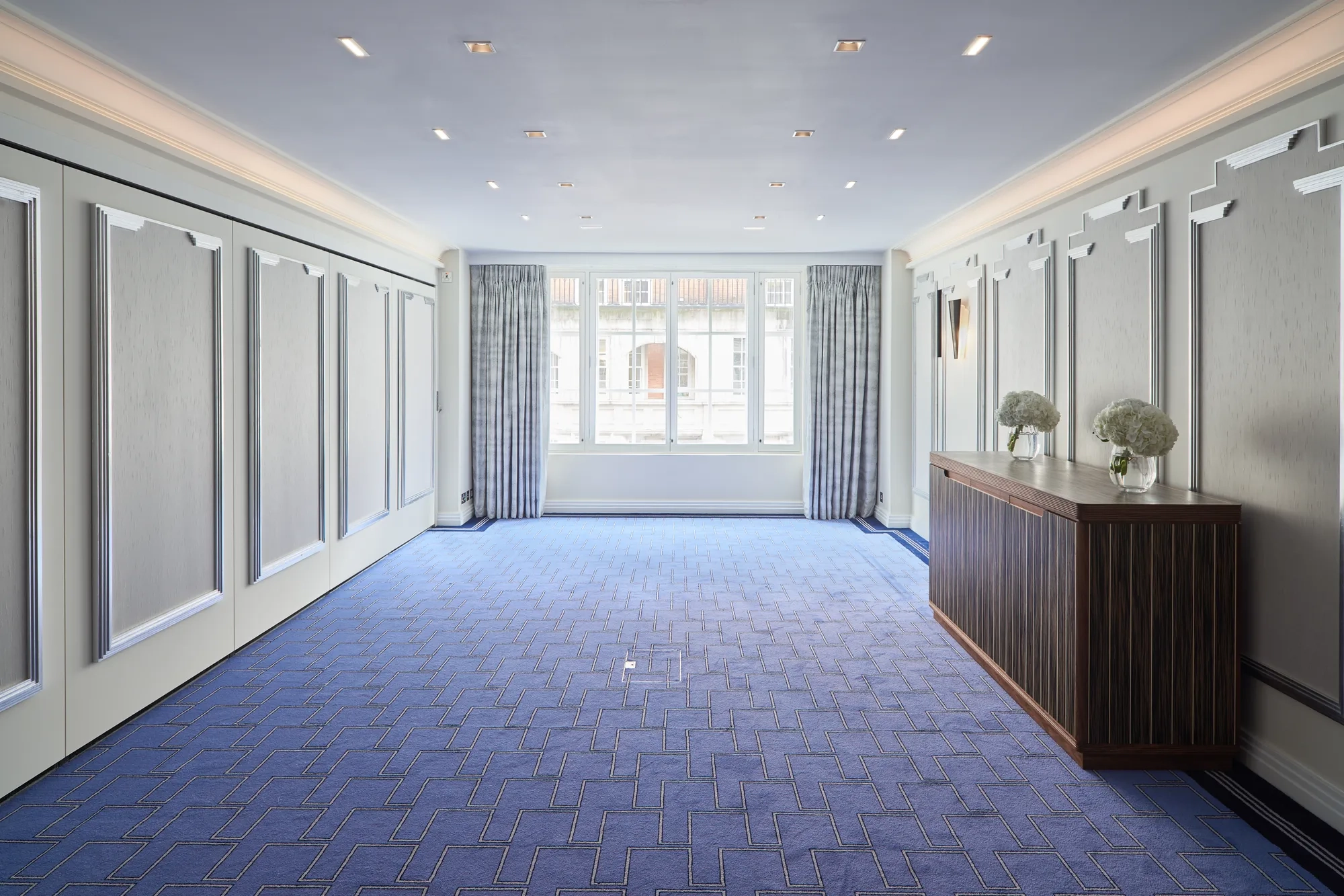 Bright, modern event room at Claridge’s featuring blue patterned carpet, soft grey panelled walls, large curtained windows, and a wooden sideboard decorated with white floral arrangements.