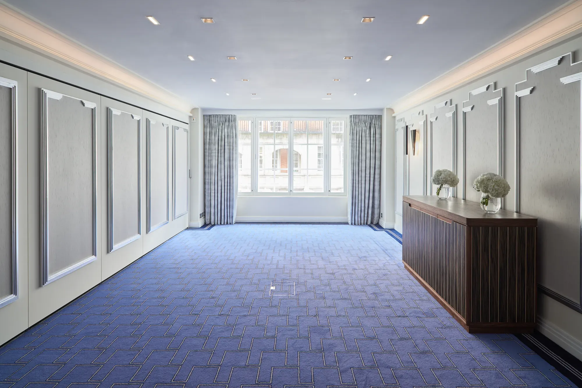 Bright, modern event room at Claridge’s featuring blue patterned carpet, soft grey panelled walls, large curtained windows, and a wooden sideboard decorated with white floral arrangements.