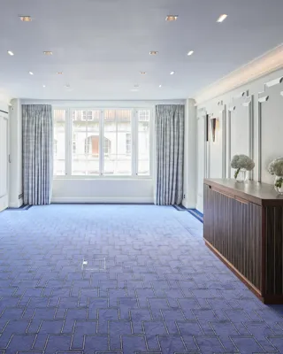 Bright, modern event room at Claridge’s featuring blue patterned carpet, soft grey panelled walls, large curtained windows, and a wooden sideboard decorated with white floral arrangements.