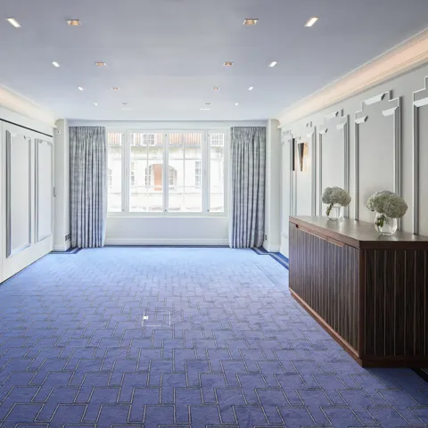 Bright, modern event room at Claridge’s featuring blue patterned carpet, soft grey panelled walls, large curtained windows, and a wooden sideboard decorated with white floral arrangements.