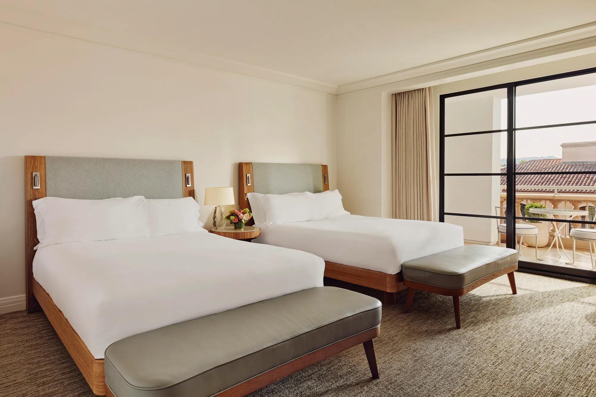 Two-queen bedroom with upholstered headboards, benches at the foot, nightstand with lamp and flowers, and doors to a balcony.