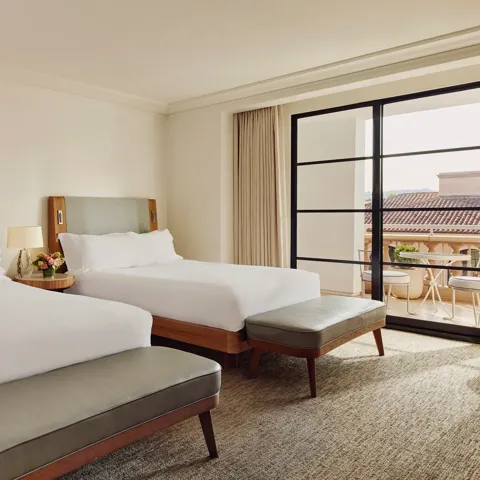 Two-queen bedroom with upholstered headboards, benches at the foot, nightstand with lamp and flowers, and doors to a balcony.