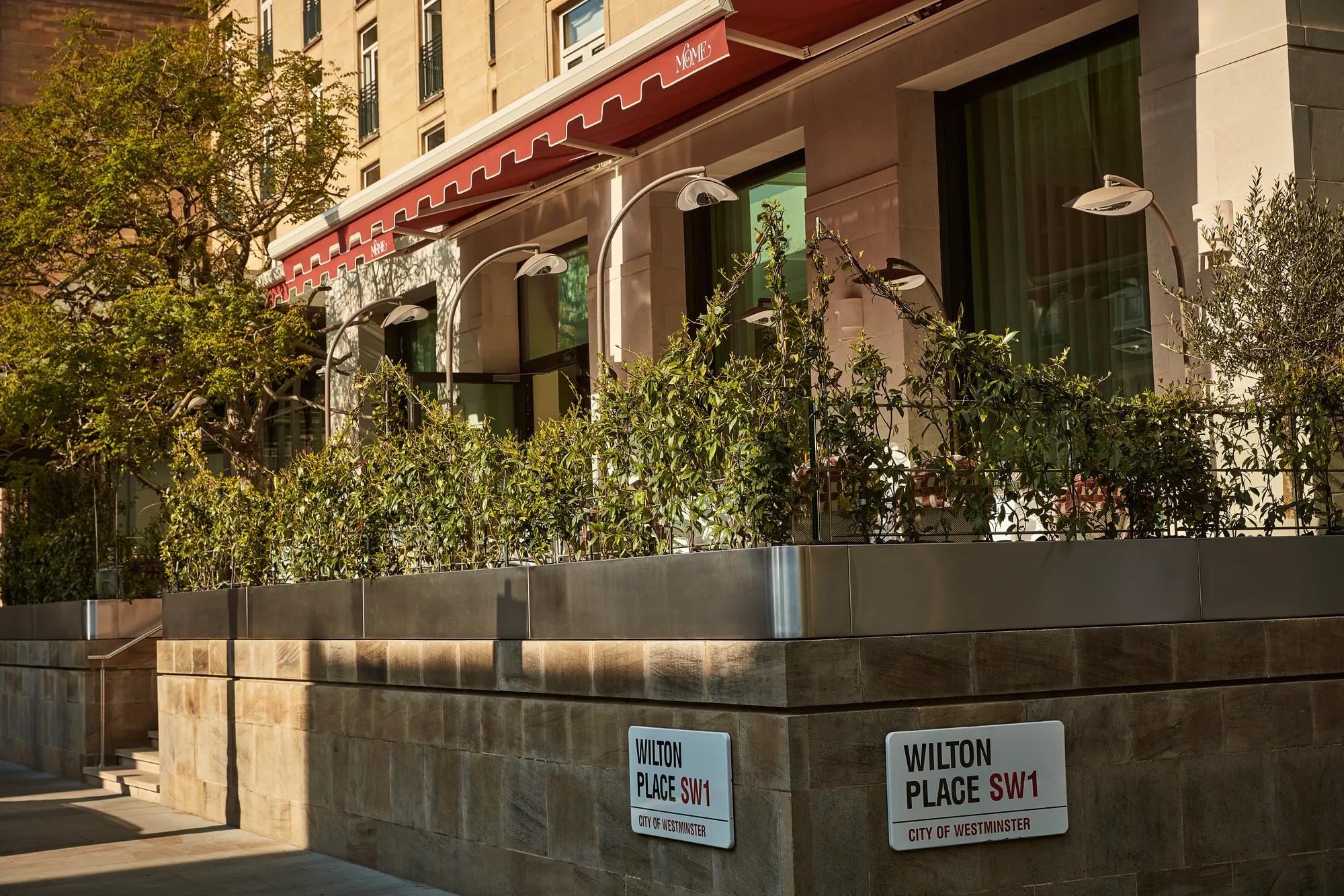 Street view of a building with a red awning and greenery along a raised terrace on Wilton Place in Westminster.