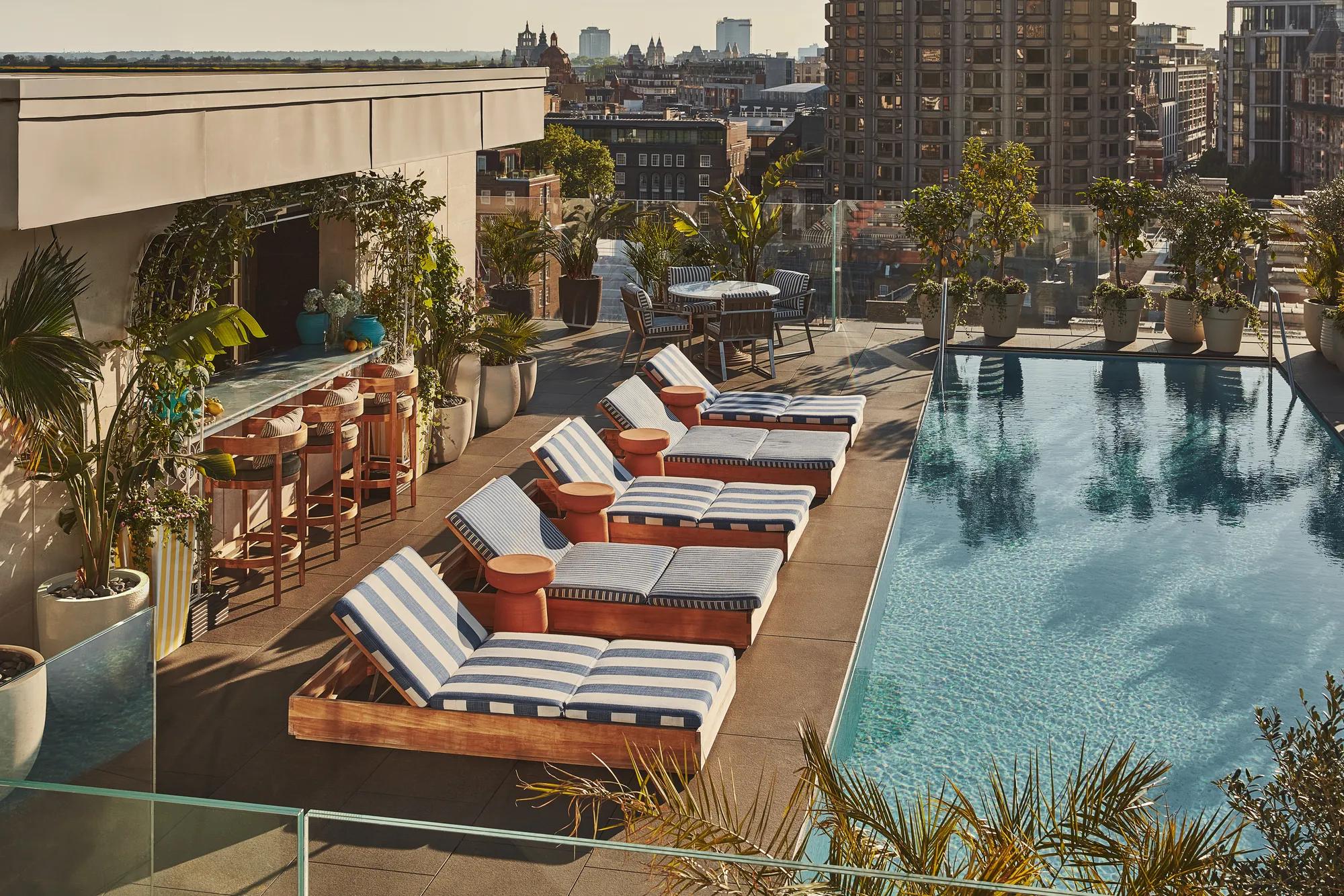 Rooftop pool with striped loungers, bar seating, greenery, and city skyline views.