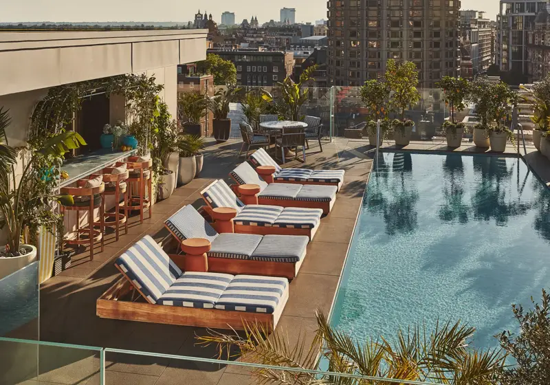 Rooftop pool with striped loungers, bar seating, greenery, and city skyline views.