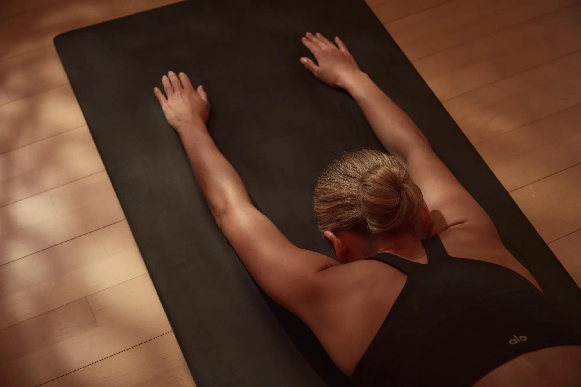 Person performing a stretching or yoga pose on a black mat, with arms extended forward and lying on their stomach, viewed from above.