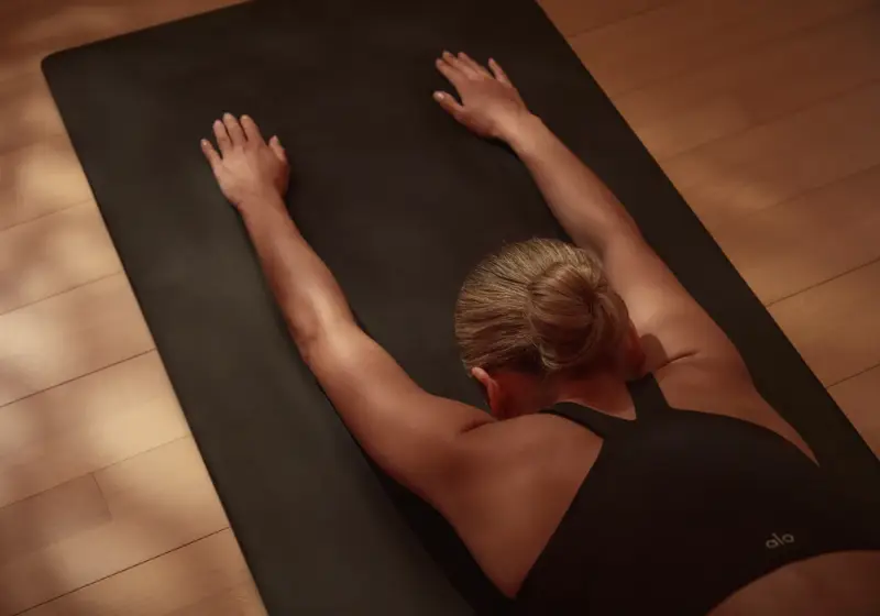 Person performing a stretching or yoga pose on a black mat, with arms extended forward and lying on their stomach, viewed from above.