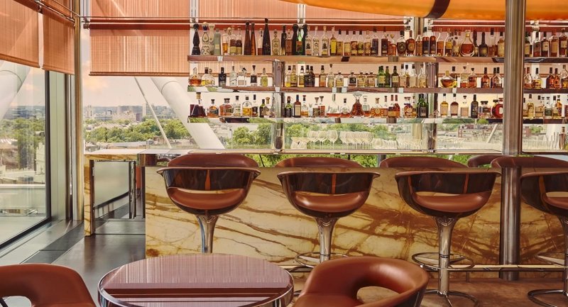 A stylish bar with marble counters, leather stools, and a panoramic city view through floor-to-ceiling windows.