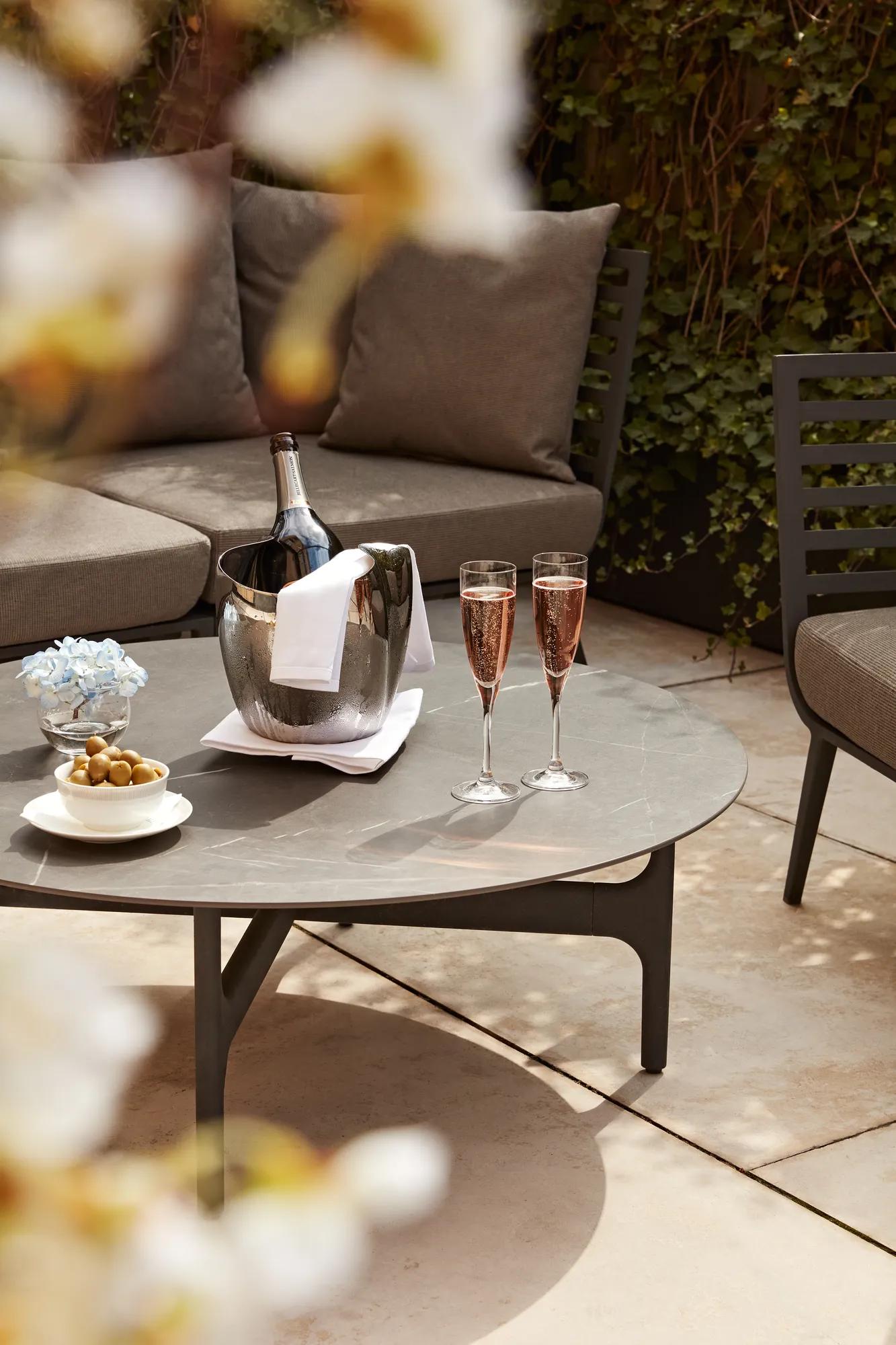Close-up of a terrace table set with a champagne bucket, two glasses of rosé, olives, and flowers, with cushioned seating behind.