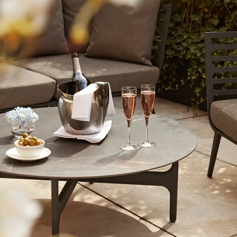 Close-up of a terrace table set with a champagne bucket, two glasses of rosé, olives, and flowers, with cushioned seating behind.