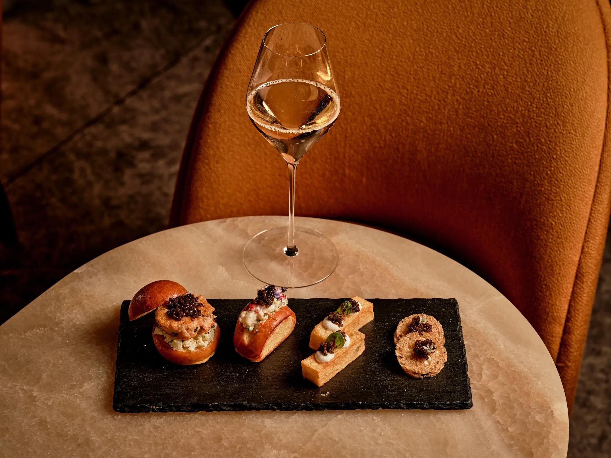 Glass of sparkling wine beside assorted caviar canapés on a black slate platter atop a marble table in a warm, elegant lounge.