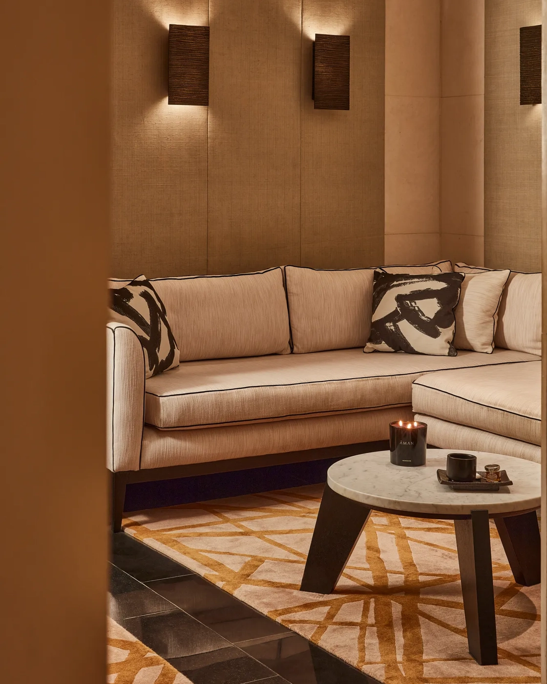 Warm lounge corner with a cream sectional sofa, abstract patterned cushions, and a low round table with a candle.