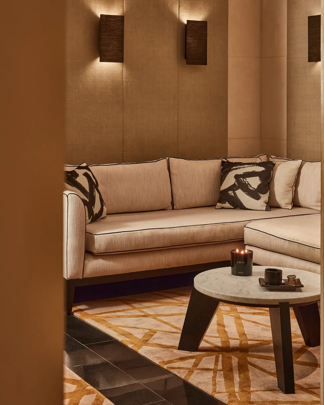 Warm lounge corner with a cream sectional sofa, abstract patterned cushions, and a low round table with a candle.