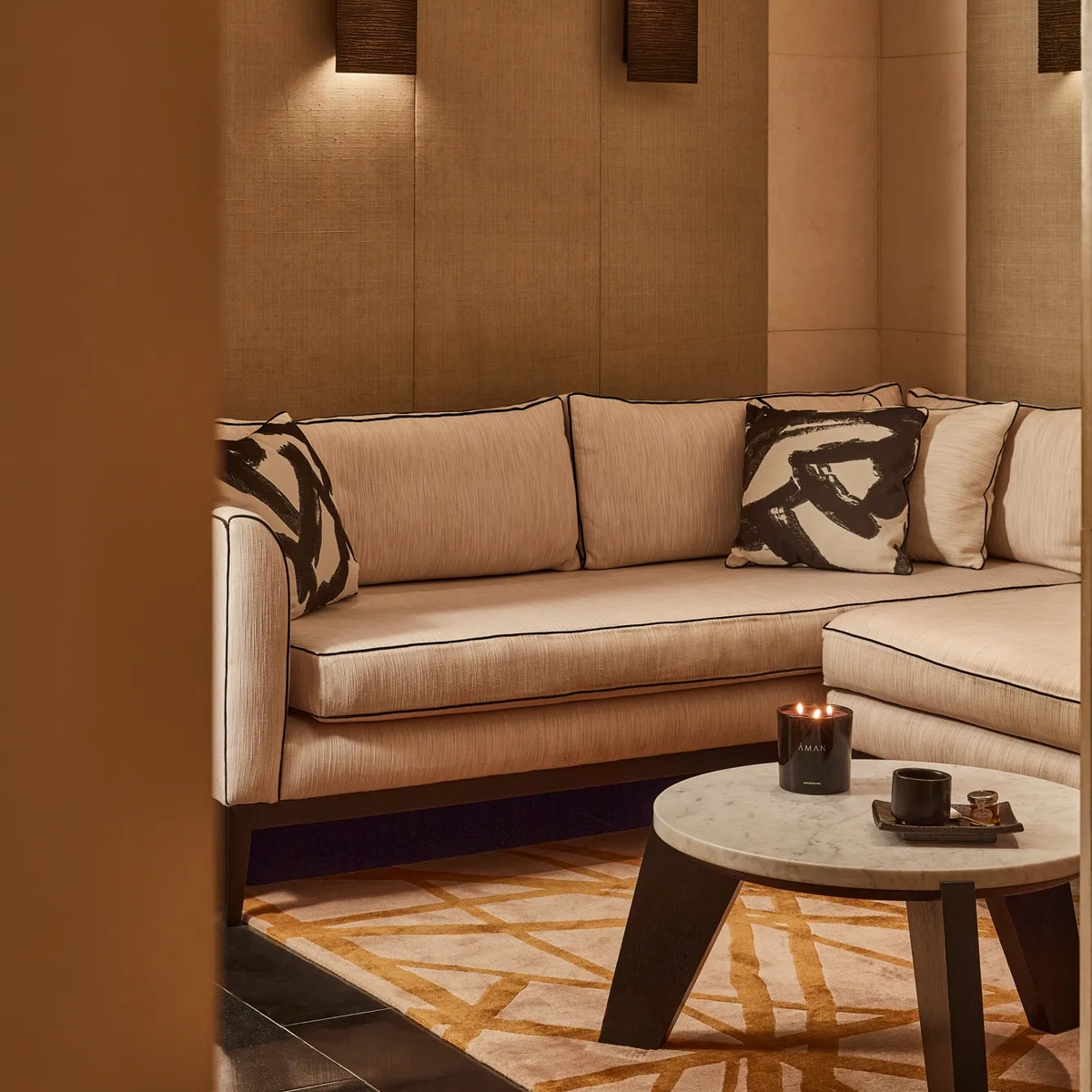 Warm lounge corner with a cream sectional sofa, abstract patterned cushions, and a low round table with a candle.