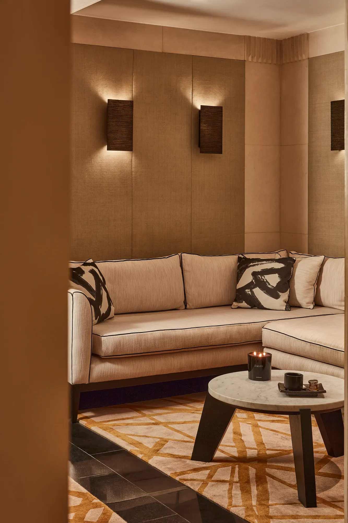 Warm lounge corner with a cream sectional sofa, abstract patterned cushions, and a low round table with a candle.