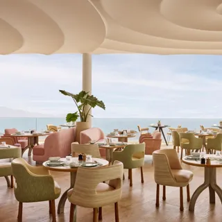 Elegant restaurant with pastel-colored chairs and round tables set with fine tableware, overlooking the sea through floor-to-ceiling windows.