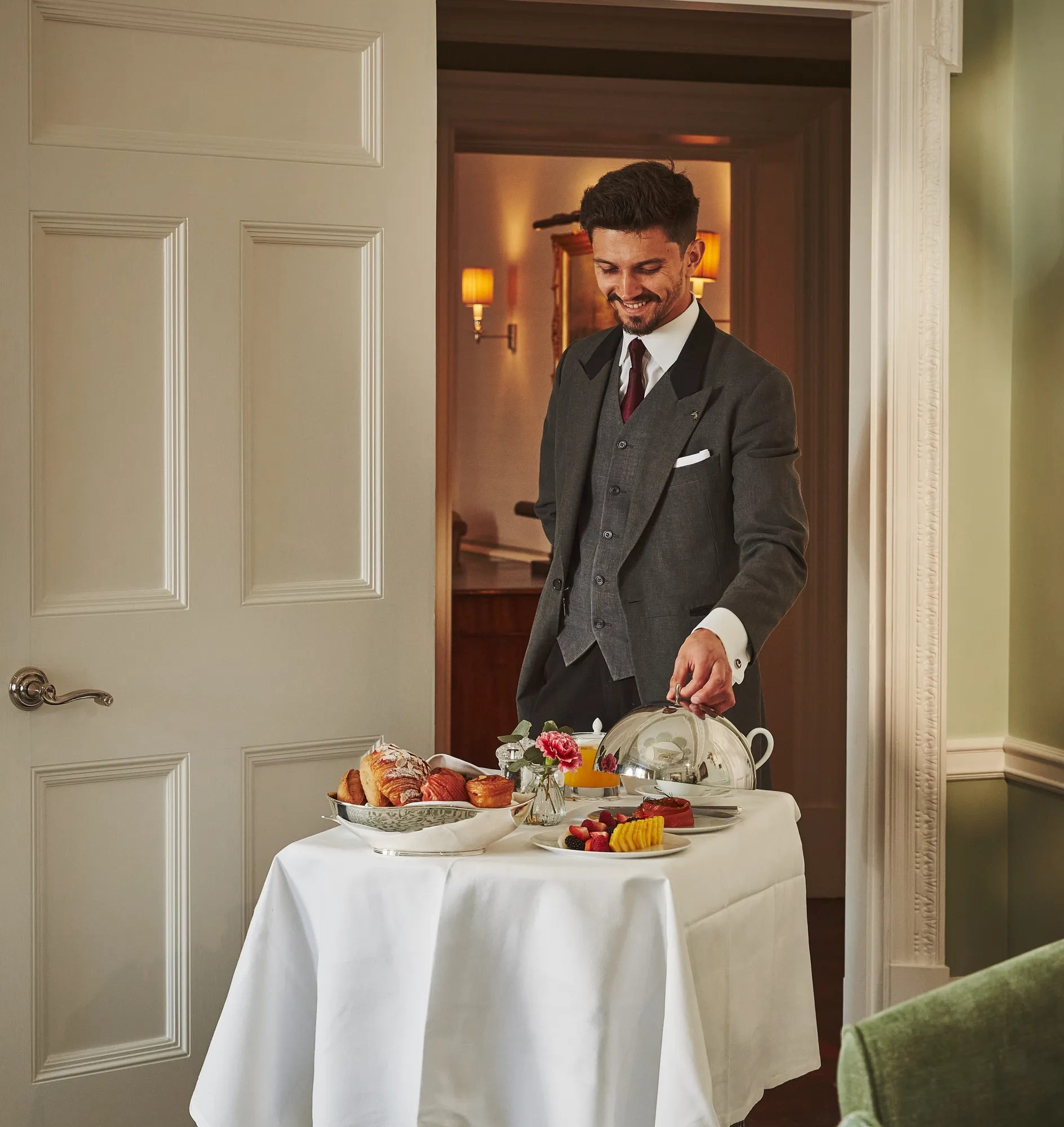 Server in a tailored suit presenting a breakfast trolley with pastries, fruit, and covered dishes in a softly lit room.