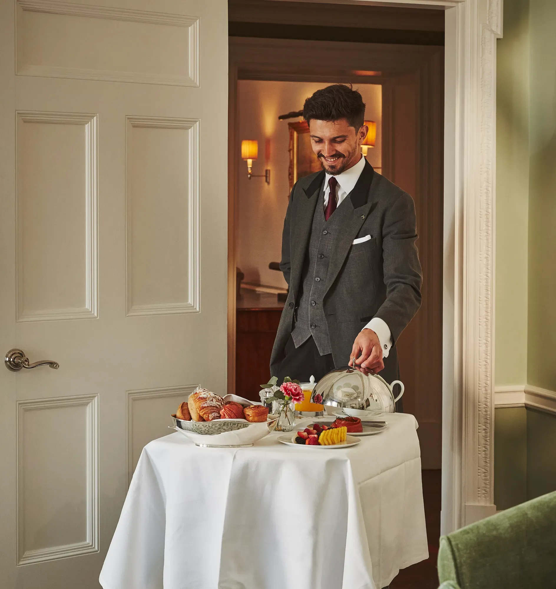 Server in a tailored suit presenting a breakfast trolley with pastries, fruit, and covered dishes in a softly lit room.