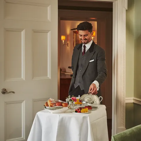Server in a tailored suit presenting a breakfast trolley with pastries, fruit, and covered dishes in a softly lit room.
