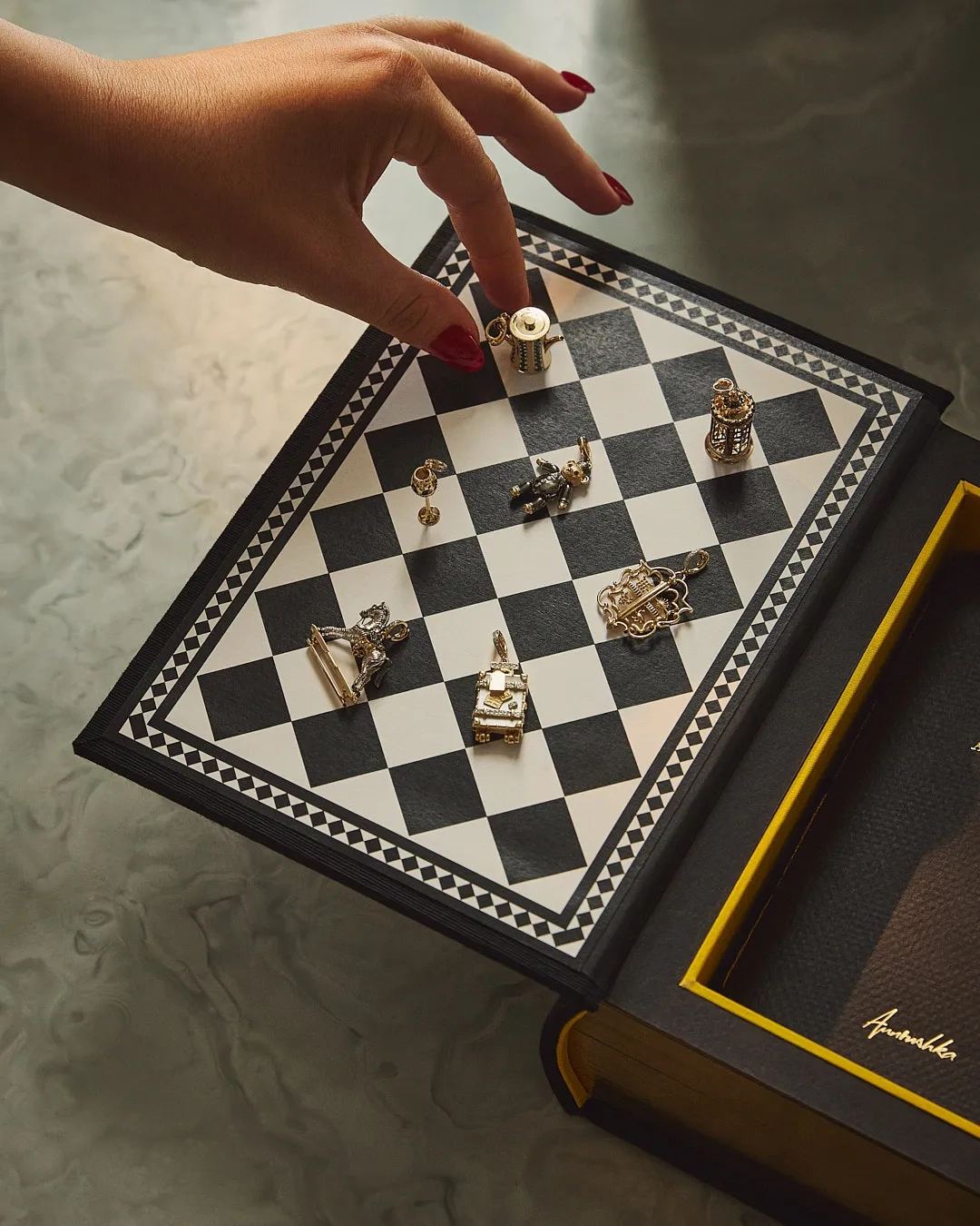 A hand with red-painted nails reaches toward a miniature chessboard embedded in a hollowed-out book, with each chess piece designed as a detailed metallic charm or figurine.