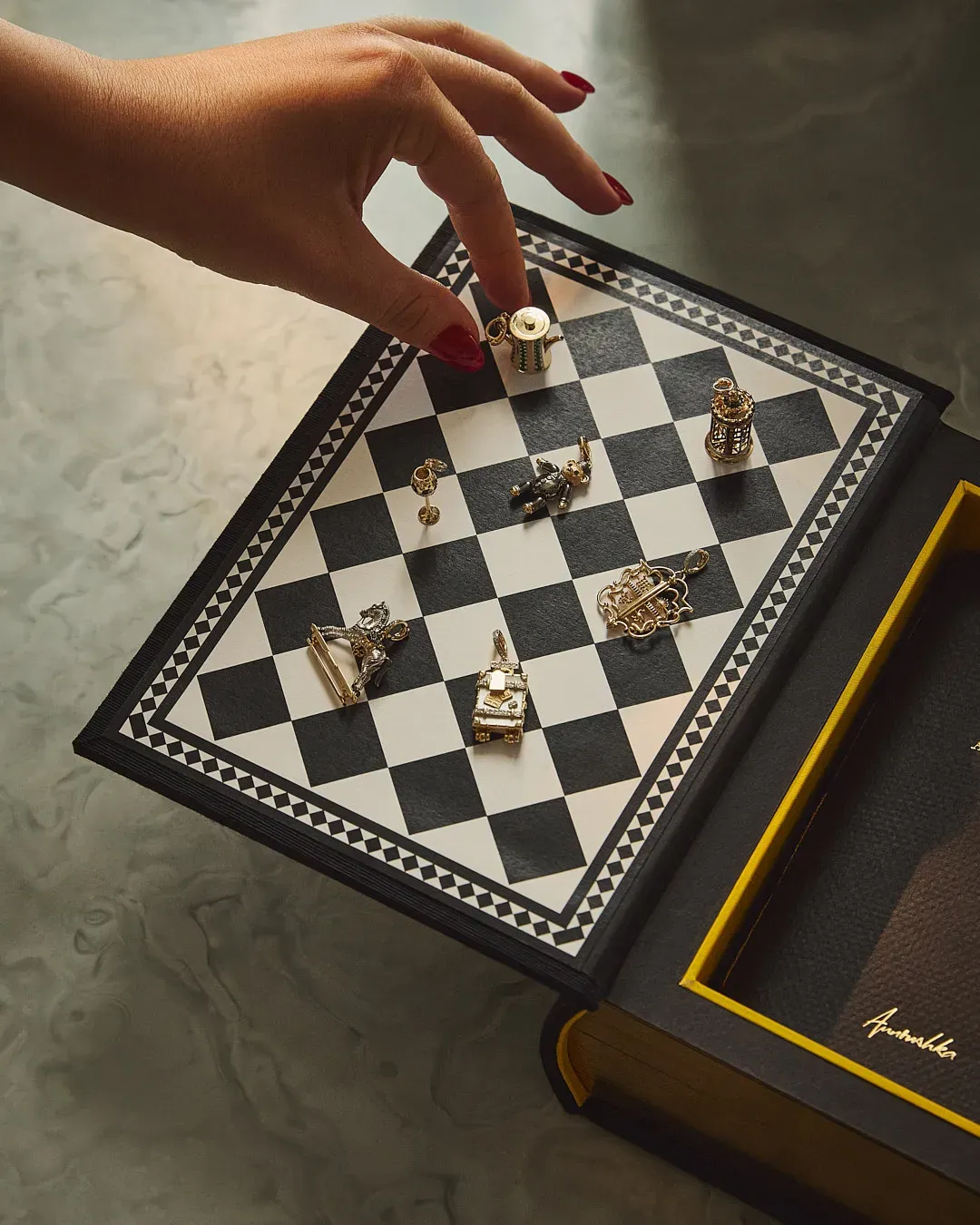 A hand with red-painted nails reaches toward a miniature chessboard embedded in a hollowed-out book, with each chess piece designed as a detailed metallic charm or figurine.