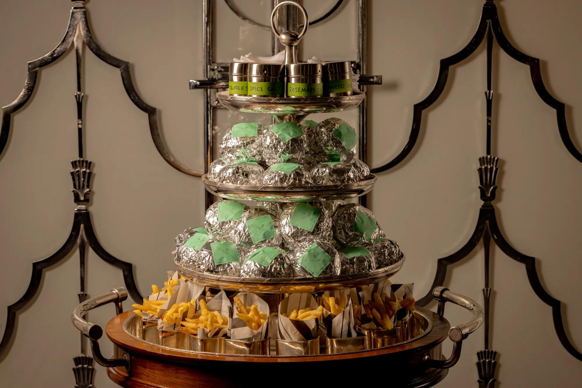 Three-tiered serving trolley at Claridge’s displaying wrapped burgers, small tins of seasoning, and portions of fries arranged neatly on a polished stand.