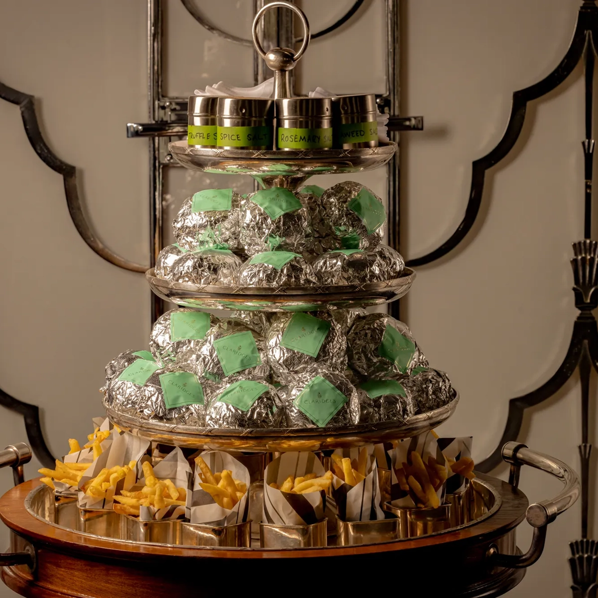 Three-tiered serving trolley at Claridge’s displaying wrapped burgers, small tins of seasoning, and portions of fries arranged neatly on a polished stand.