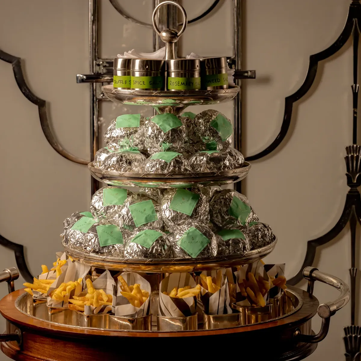 Three-tiered serving trolley at Claridge’s displaying wrapped burgers, small tins of seasoning, and portions of fries arranged neatly on a polished stand.