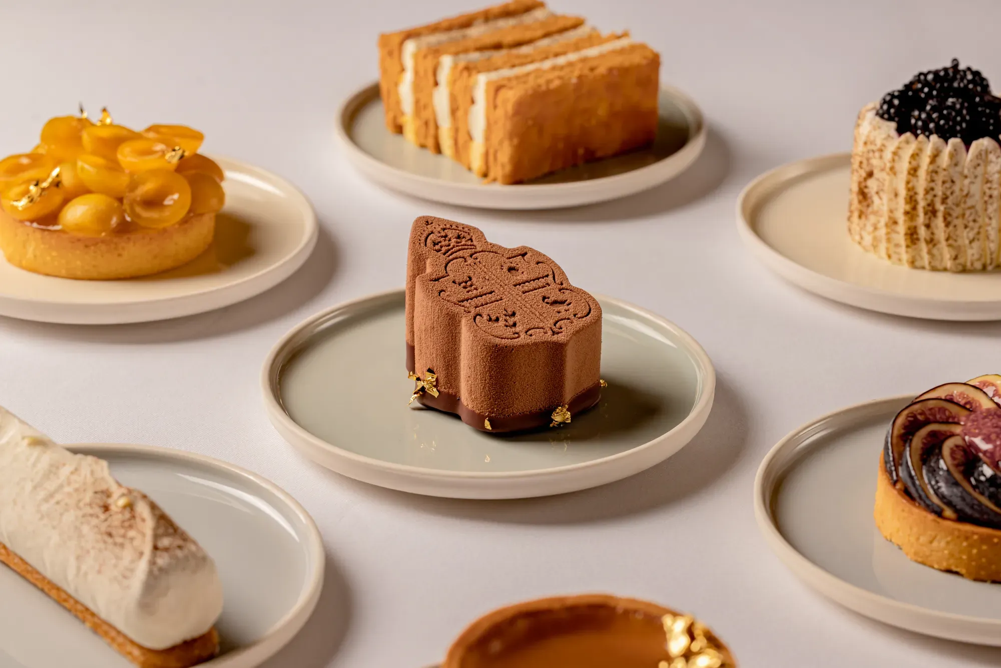 Selection of elegant pastries on neutral plates, including a chocolate mousse dessert with embossed design, fruit tarts, mille-feuille, and cream-filled cakes, arranged on a light surface.