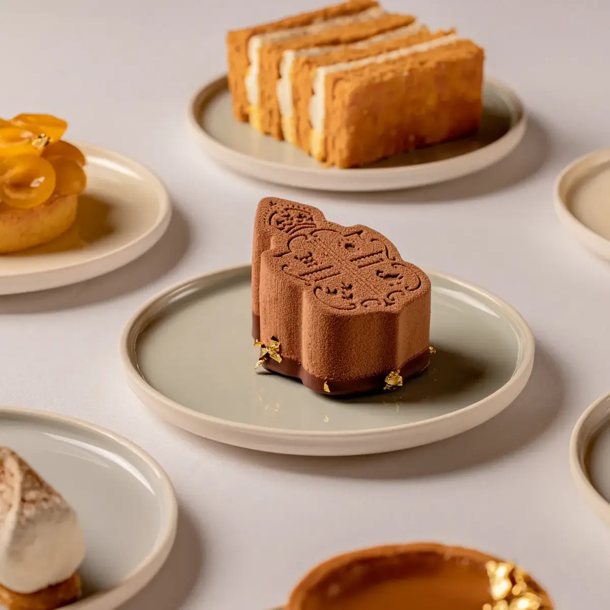 Selection of elegant pastries on neutral plates, including a chocolate mousse dessert with embossed design, fruit tarts, mille-feuille, and cream-filled cakes, arranged on a light surface.