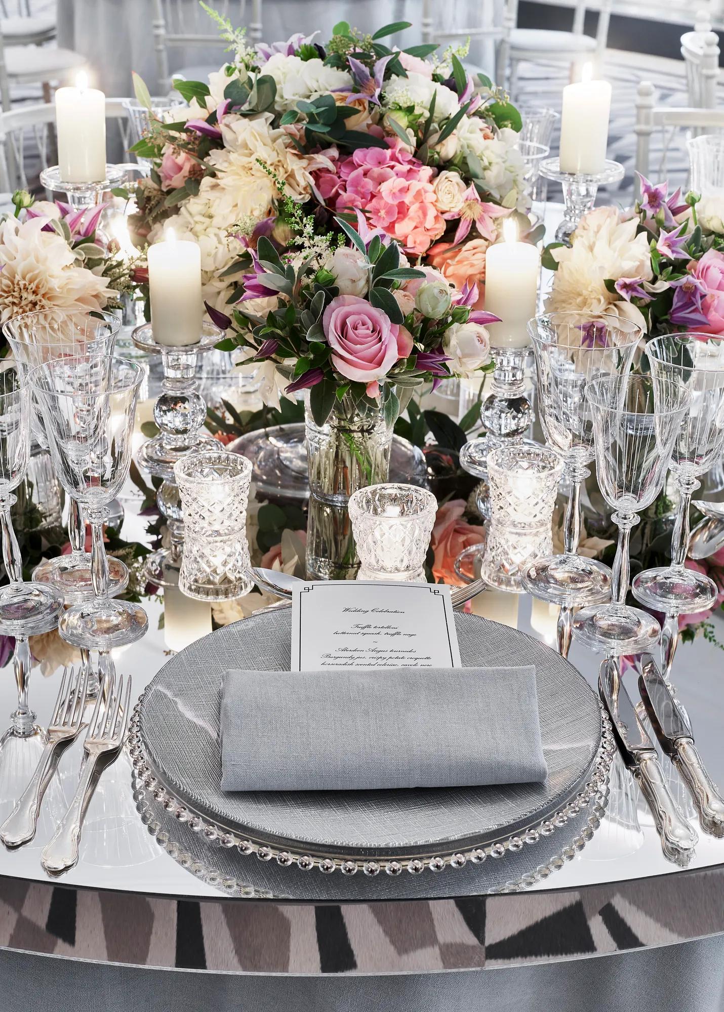 Elegant place setting with floral centerpiece, candles, and crystal glassware on mirrored table.