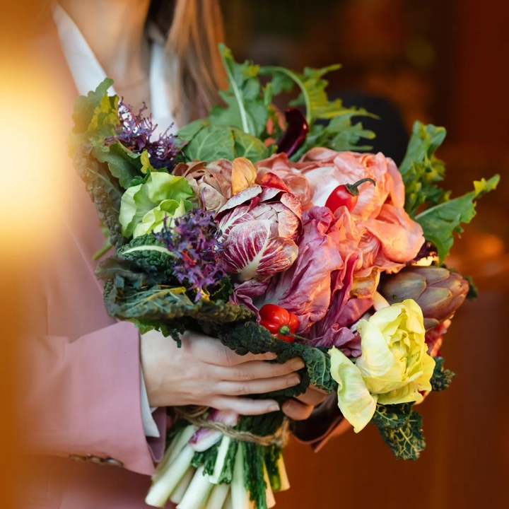 Person in a pink blazer holding a bouquet of leafy greens, cabbage, artichoke and radicchio in warm light.