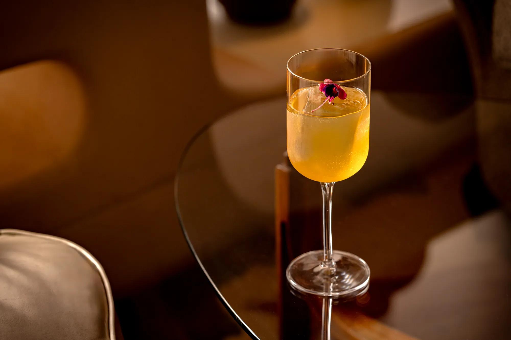 Golden cocktail with flower garnish in stemmed glass on dark wood table.